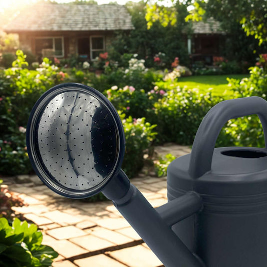 Gros plan sur la pomme d'un arrosoir gris foncé, avec un jardin fleuri et une maison de campagne en arrière-plan sous un soleil éclatant.