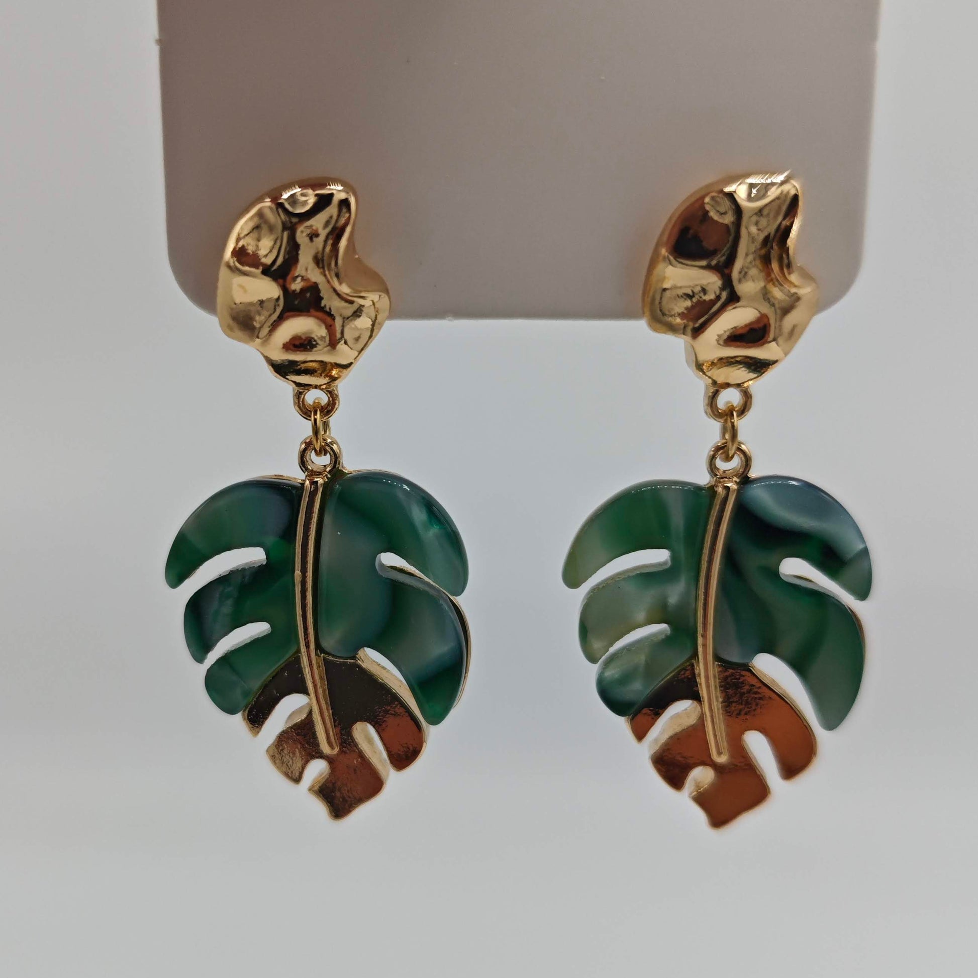 Une paire de boucles d'oreilles pendantes en forme de feuilles de Monstera, de couleur or et verte, présentées sur un support blanc.
