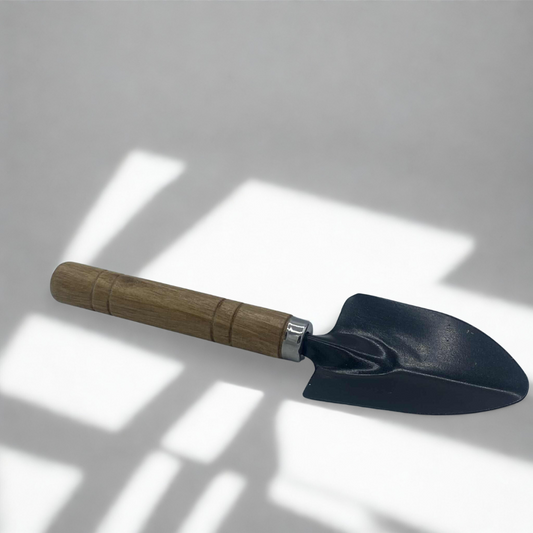 Une petite pelle de jardinage avec une tête en métal noir et un manche en bois clair, posée sur un fond blanc avec des ombres.