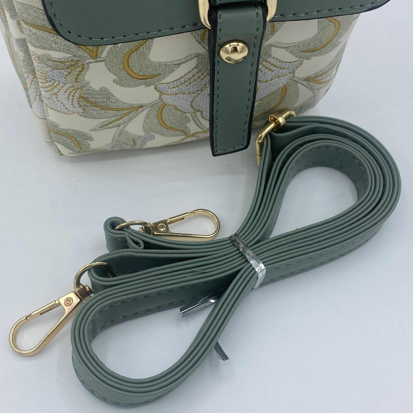 Gros plan sur la bandoulière de couleur vert sauge et les mousquetons dorés d'un sac orné d'un motif de feuilles brodé.