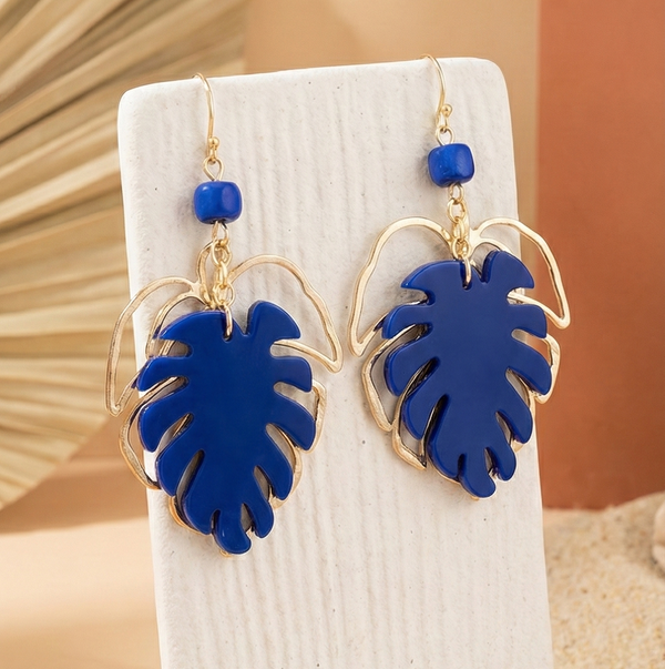 Paire de boucles d'oreilles pendantes en forme de feuille de monstera, combinant une partie pleine en acrylique bleu vif et une monture ajourée en métal doré. Elles sont présentées sur un support clair dans un décor aux tons naturels.