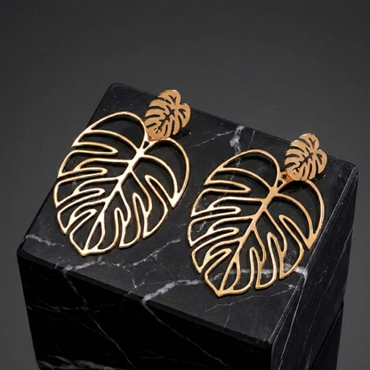 Gros plan sur des boucles d'oreilles motif feuille de Monstera en métal doré, posées sur un cube de marbre noir veiné, offrant un contraste élégant et sophistiqué.