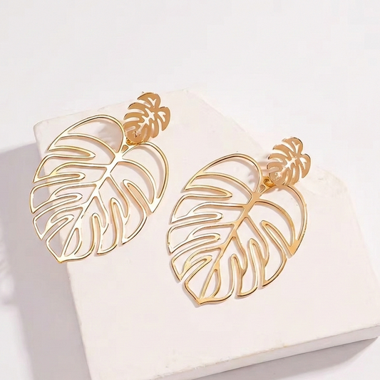 Paire de boucles d'oreilles dorées en forme de feuille de Monstera au design ajouré, présentées à plat sur un support en pierre blanche texturée avec un éclairage doux et naturel.