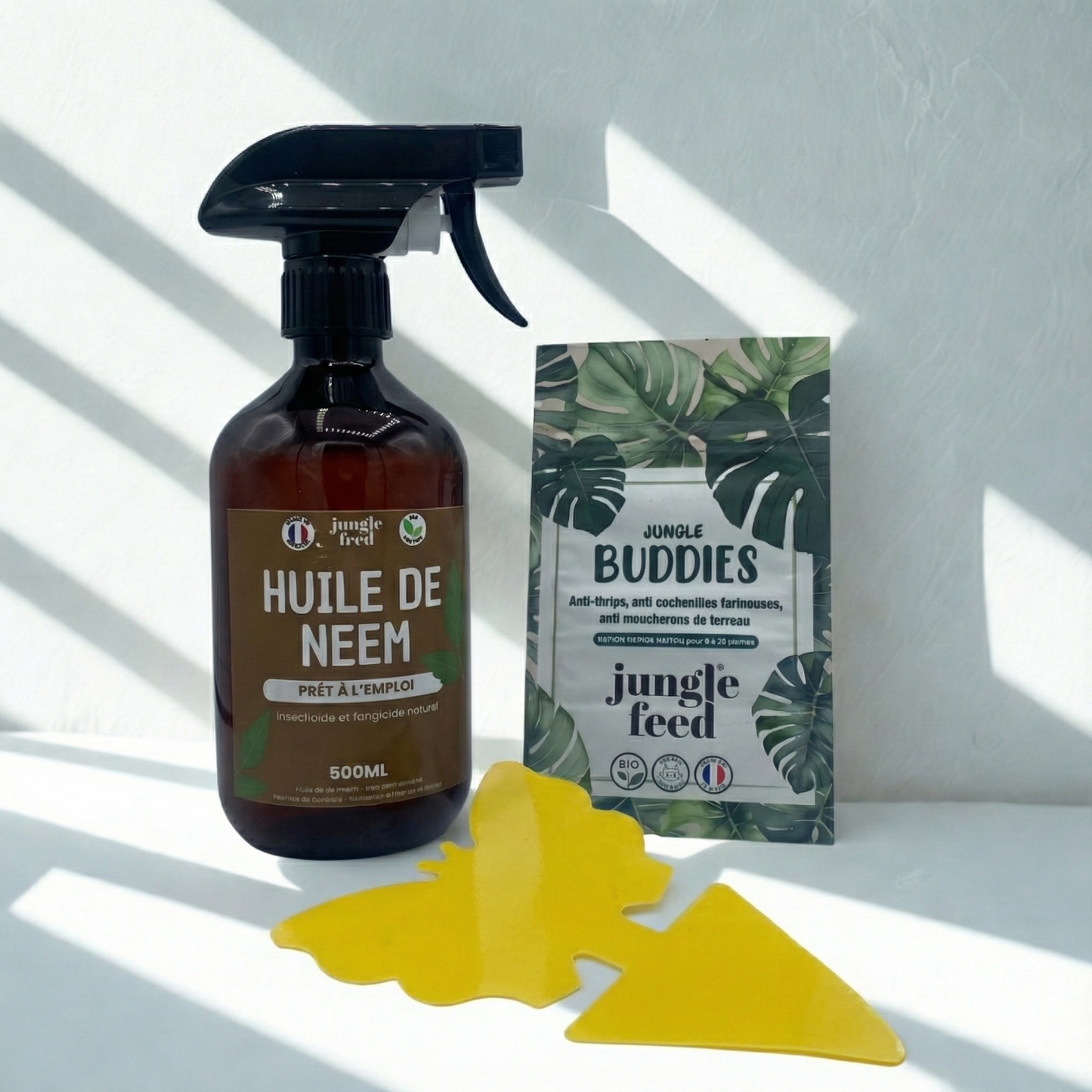 Plan rapproché sur un kit d'entretien pour plantes d'intérieur Jungle Feed comprenant un spray d'huile de Neem et un sachet de protection naturelle. Un piège à insectes bleu en forme de flèche et un piège papillon sont disposés devant les produits, sous une lumière naturelle créant des ombres rayées.