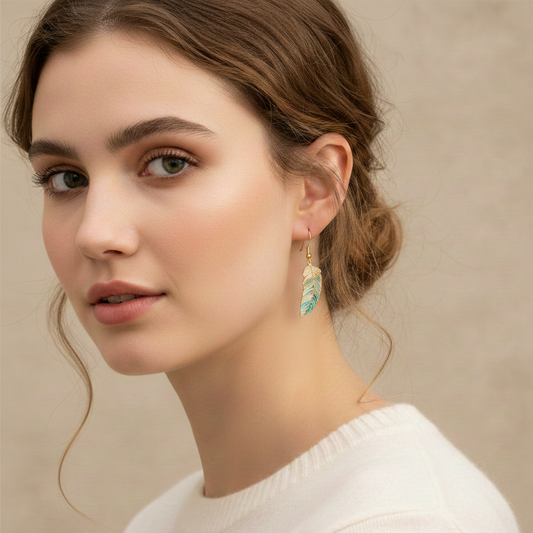 Portrait de profil d'une femme portant une boucle d'oreille pendante en forme de plume aux reflets bleu sarcelle et dorés. Elle porte un pull blanc et ses cheveux sont attachés en un chignon flou.