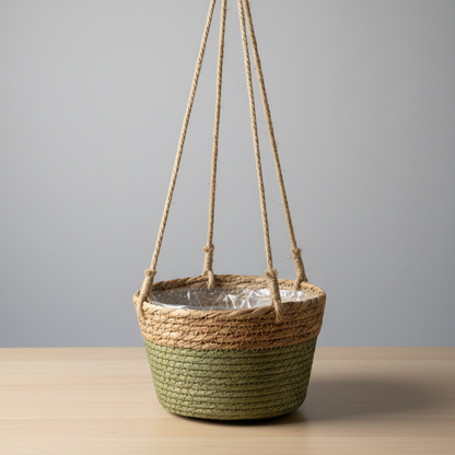 Panier tressé bicolore suspendu (vert olive et beige clair), avec une doublure en plastique transparent et trois cordes de jute le suspendant, posé sur une table en bois clair devant un mur gris uni.
