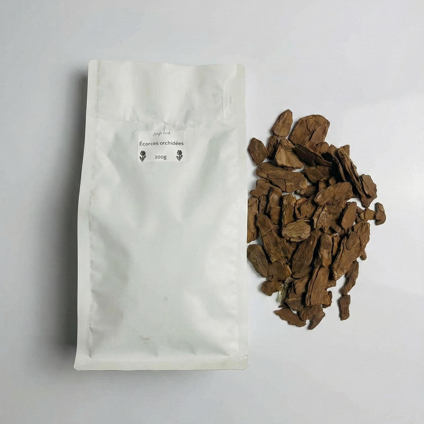 Vue de dessus d'un sachet blanc hermétique étiqueté « Écorces orchidées 200g » posé à côté d'un tas de copeaux d'écorce de pin bruns sur un fond blanc épuré.