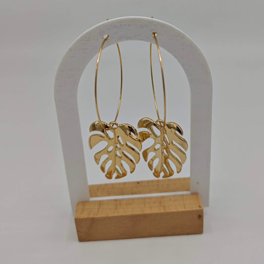 Une paire de boucles d'oreilles créoles dorées, avec des pendentifs en forme de feuilles de Monstera, présentées sur un support blanc et en bois.