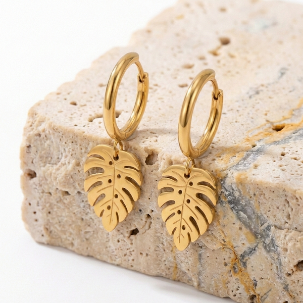 Boucles d'oreilles motif feuille tropicale monstera posées sur un support en pierre naturelle texturée beige. Mise en scène élégante mettant en valeur la brillance du métal doré.