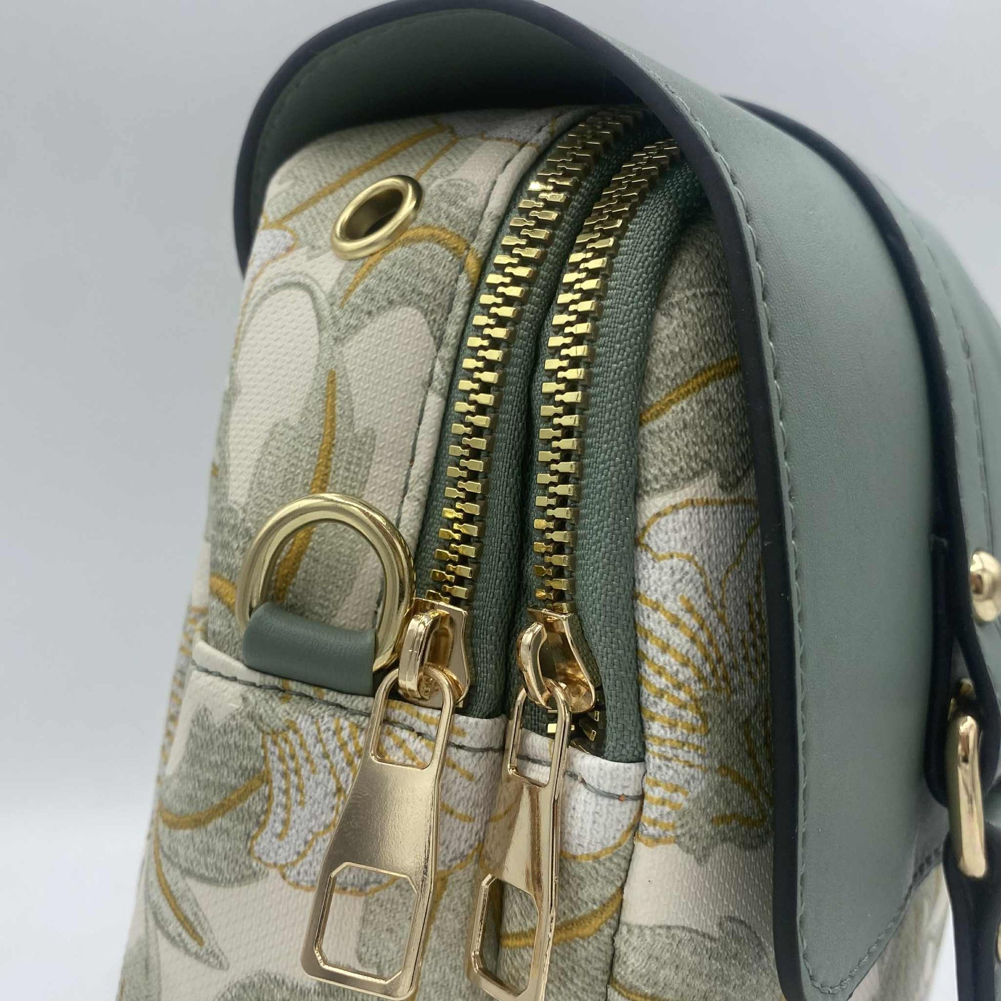 Gros plan sur le dessus d'un sac à main avec deux fermetures éclair dorées. Le sac est décoré d'un motif de feuilles vert clair sur fond blanc et d'un rabat vert sauge.