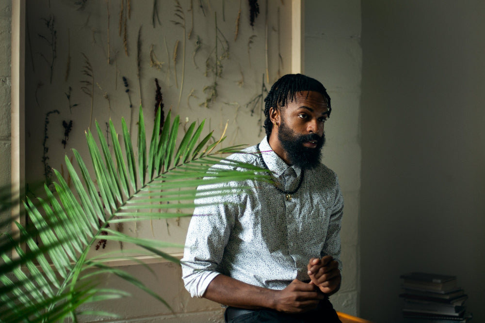 Un homme avec une barbe, habillé d'une chemise à motifs, est assis près d'une grande plante verte. Derrière lui, une œuvre murale avec des plantes séchées.
