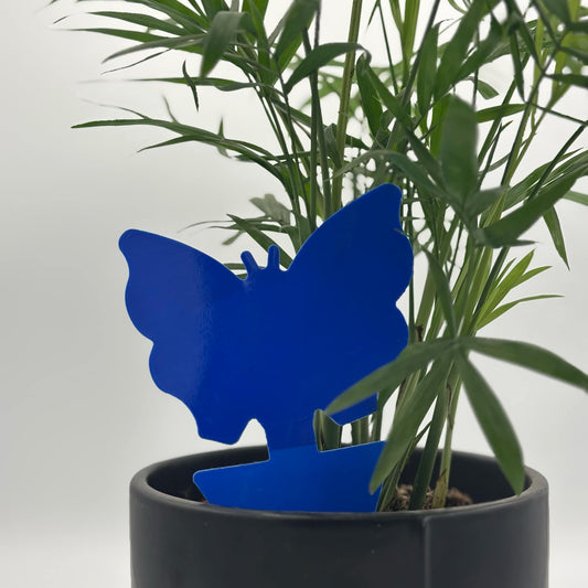 Un piège adhésif bleu en forme de papillon, inséré dans la terre d'un pot de plante verte d'intérieur. Le pot est noir et hexagonal, et l'arrière-plan est unifié et clair.