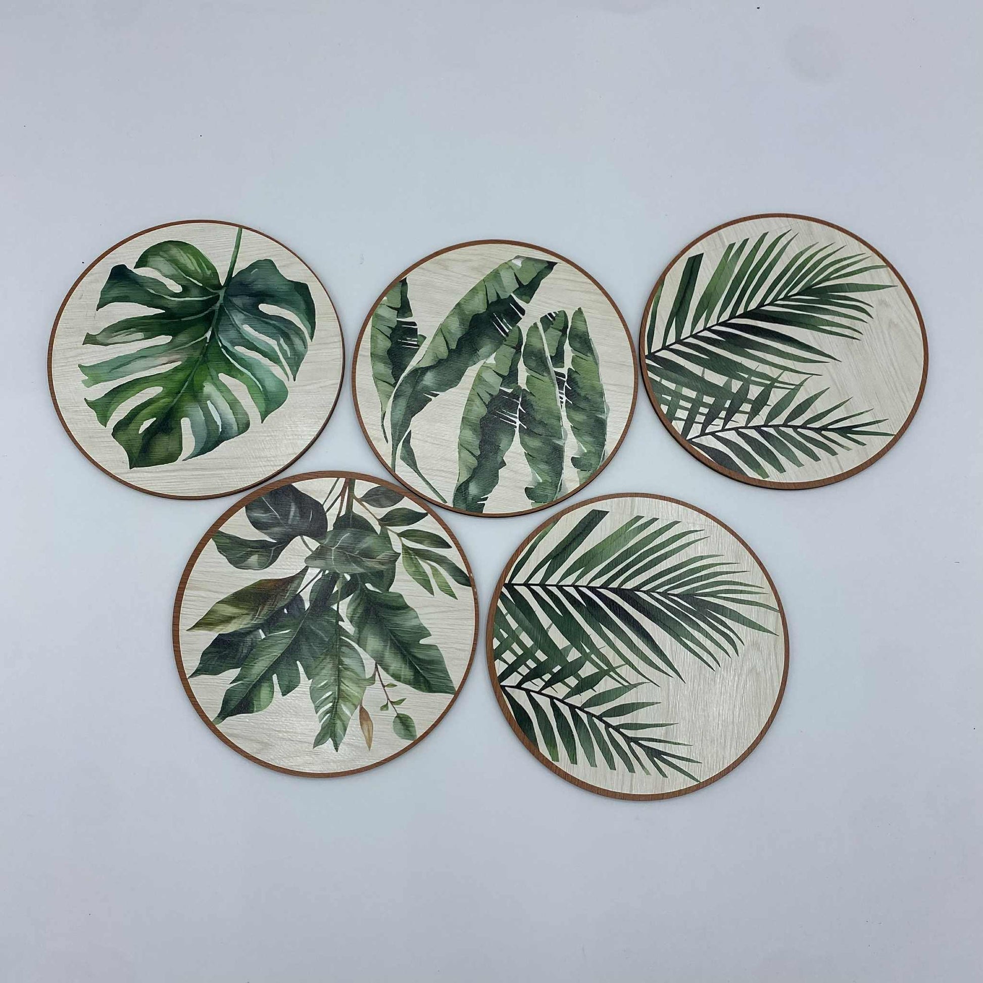  Un ensemble de cinq sous-verres ronds avec des motifs de feuilles de plantes tropicales, dont une feuille de Monstera, sur un fond effet bois clair