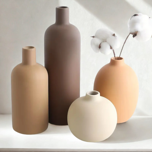 Quatre vases minimalistes en céramique de formes et de tailles différentes, dans des tons neutres comme le beige et le marron, posés sur une étagère. Le vase le plus grand contient des branches de coton.